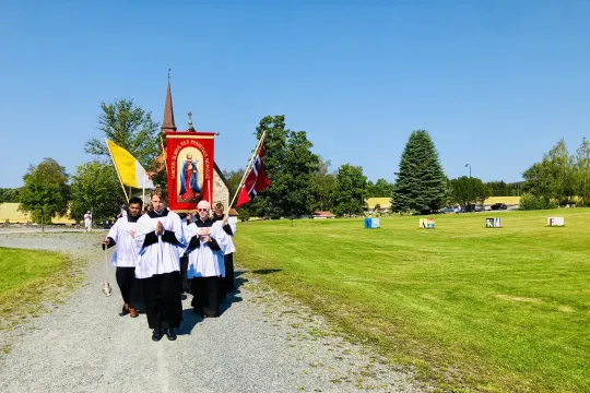 Prosesjon under olsokfeiringen på Stiklestad i 2021. Sol. Blå himmel. Gamle Stiklestad kirke i bakgrunnen.
