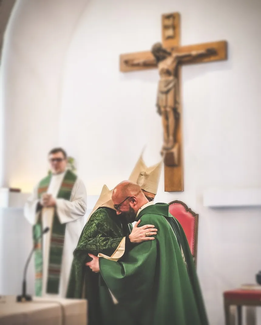 Biskop Erik Varden og pater Rafał Ochojski omfavner hverandre ved alteret i Vår Frue domkirke i Tromsø. I bakgrunnen henger et stort krusifiks.
